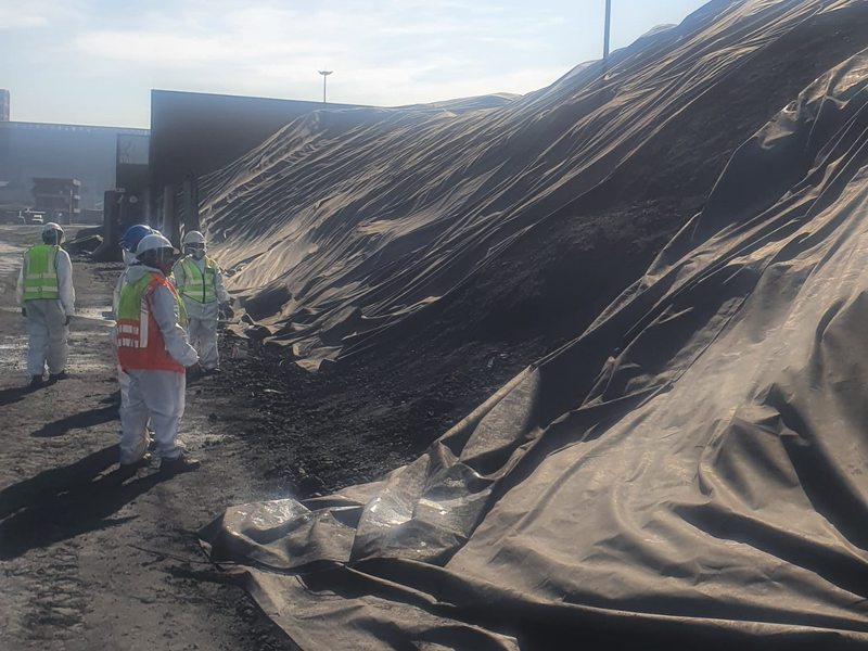 Inspectors in coveralls and hi-vis beside a long tarped coal stockpile